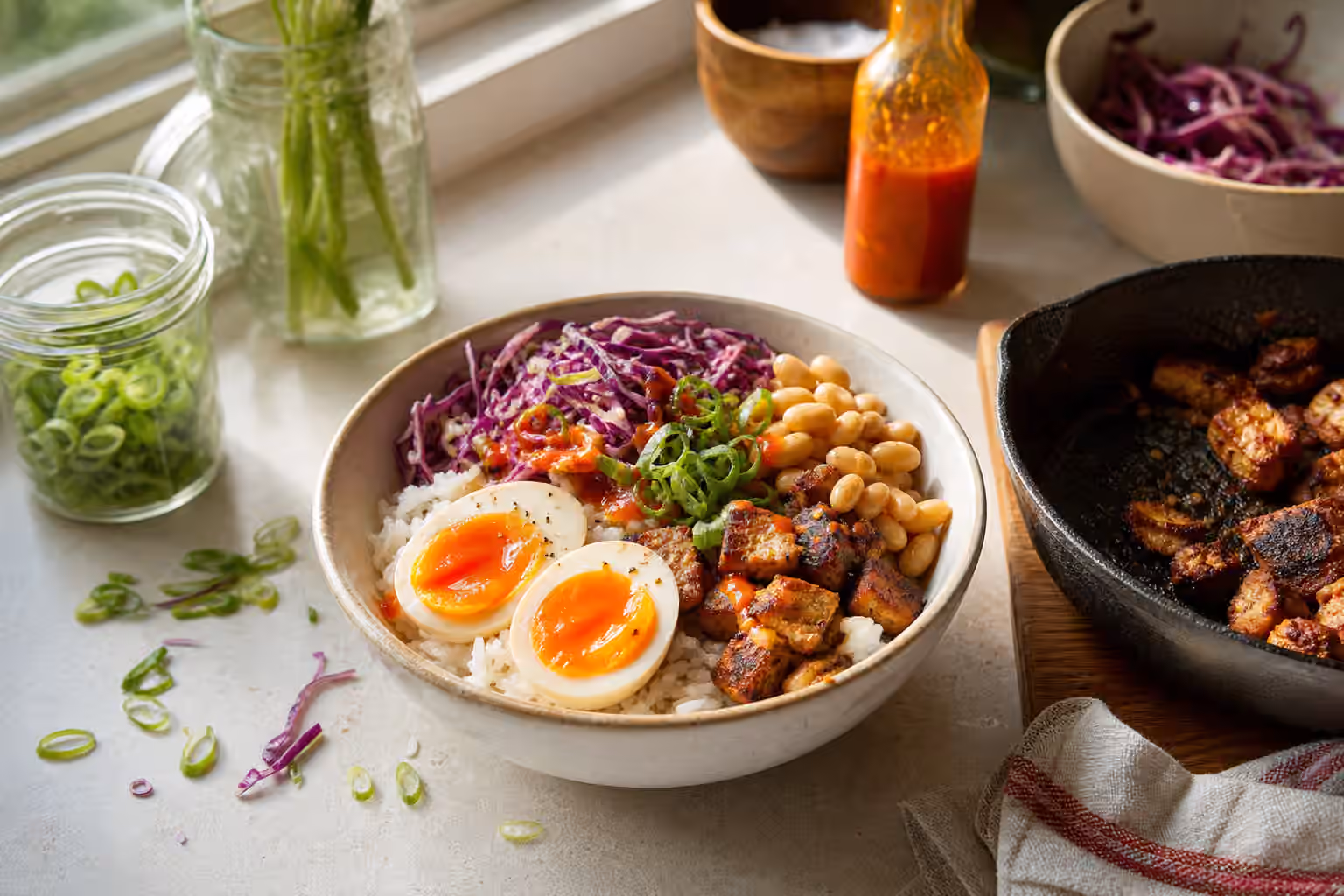 A rice bowl with jammy eggs, browned protein, beans, cabbage, scallions, and hot sauce.