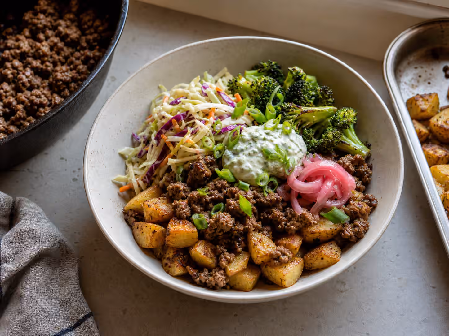A potato boy kibble bowl with roasted potatoes, browned meat, broccoli, slaw, herb sauce, and pickled onions.