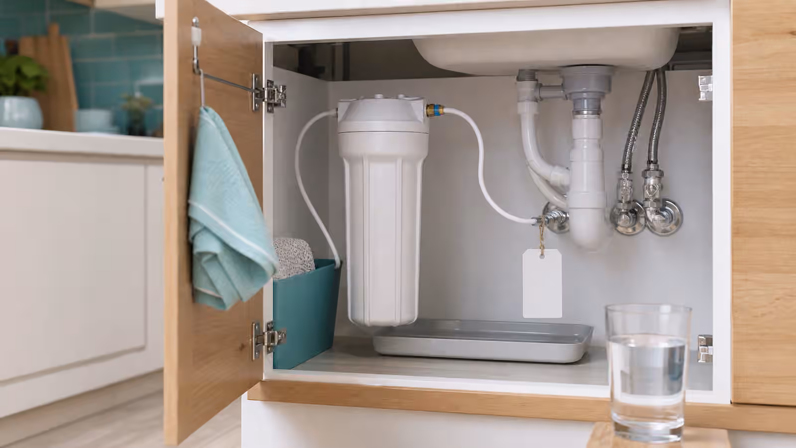 An organized under-sink cabinet with filter housing, tubing, shutoff valves, leak tray, and a glass of water.