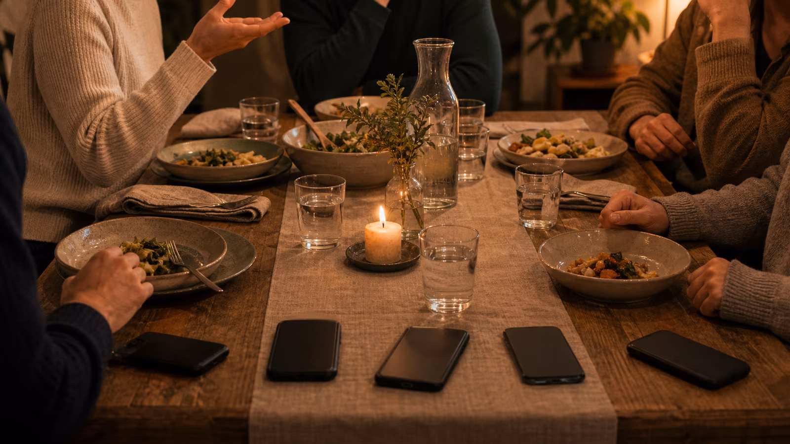 a small dinner table with phones face down near the edge, warm plates, glasses, candlelight, and people leaning into conversation