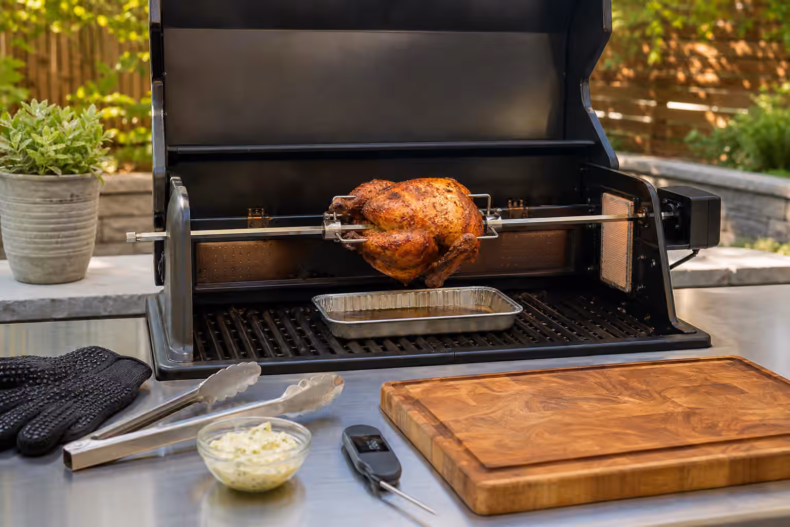 A browned chicken turning on a rotisserie spit over a drip pan in a clean backyard grill.