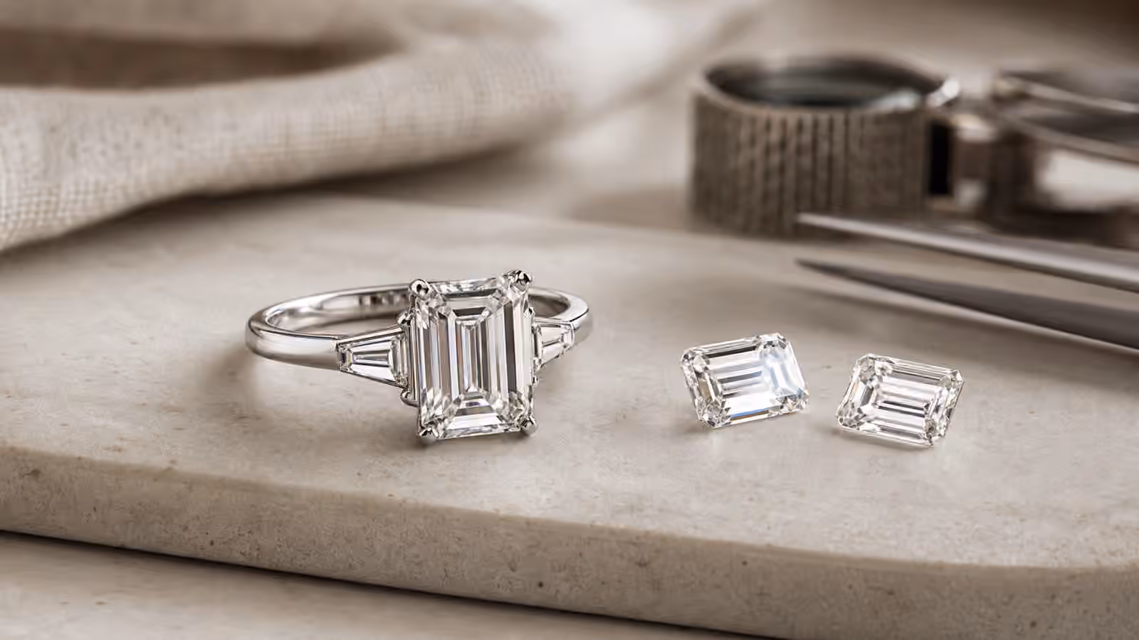 Emerald-cut engagement ring beside two loose rectangular step-cut diamonds on a jeweler's tray.