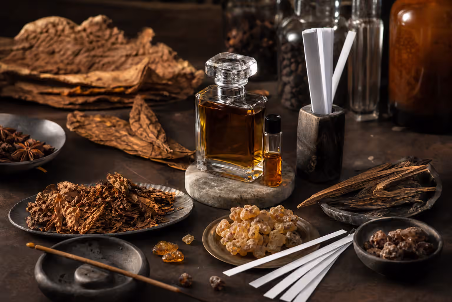 An unbranded perfume bottle with blotter strips, cured tobacco leaves, incense resin, dry woods, amber resin tears, spice pods, and a ceramic dish on a dark workbench.