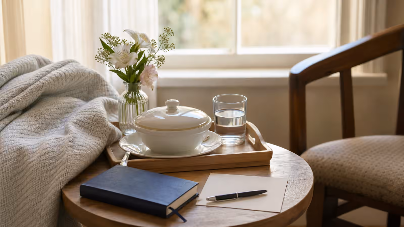 A quiet care table with covered soup, water, flowers, a closed prayer book, blank note card, pen, blanket, and empty chair.