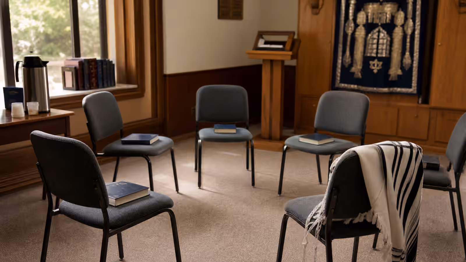 A small synagogue chapel with chairs, prayer books, a folded tallit, lectern, and morning light.