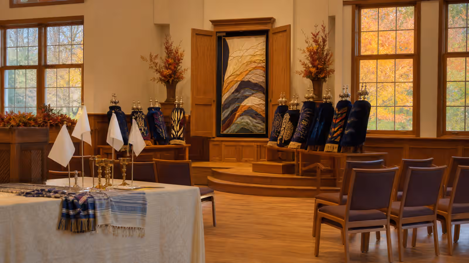 A synagogue prepared for Simchat Torah with covered Torah scrolls, open space, chairs, blank flags, and autumn light.