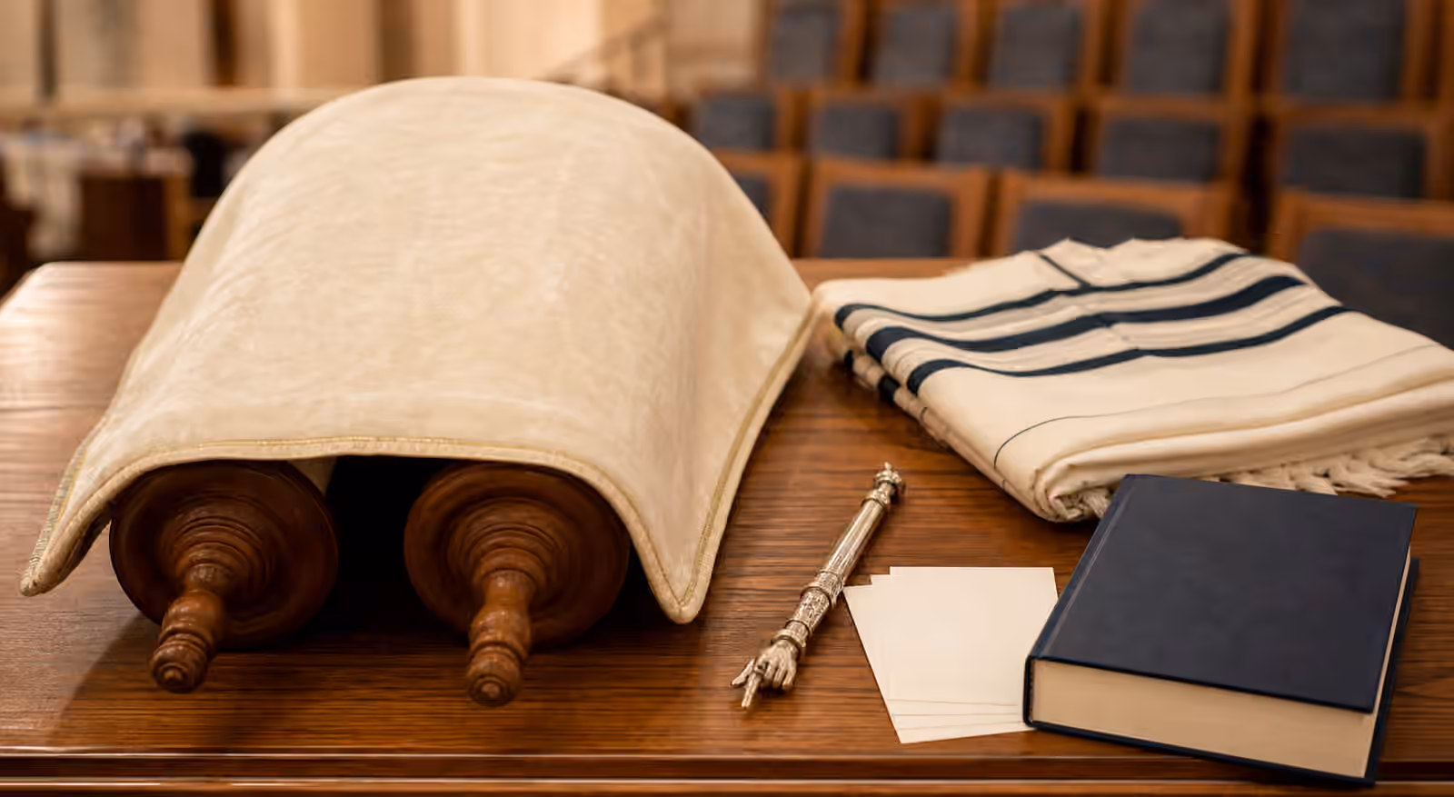 A covered Torah scroll on a wooden reading table with a yad pointer, folded tallit, blank cards, and a plain book.
