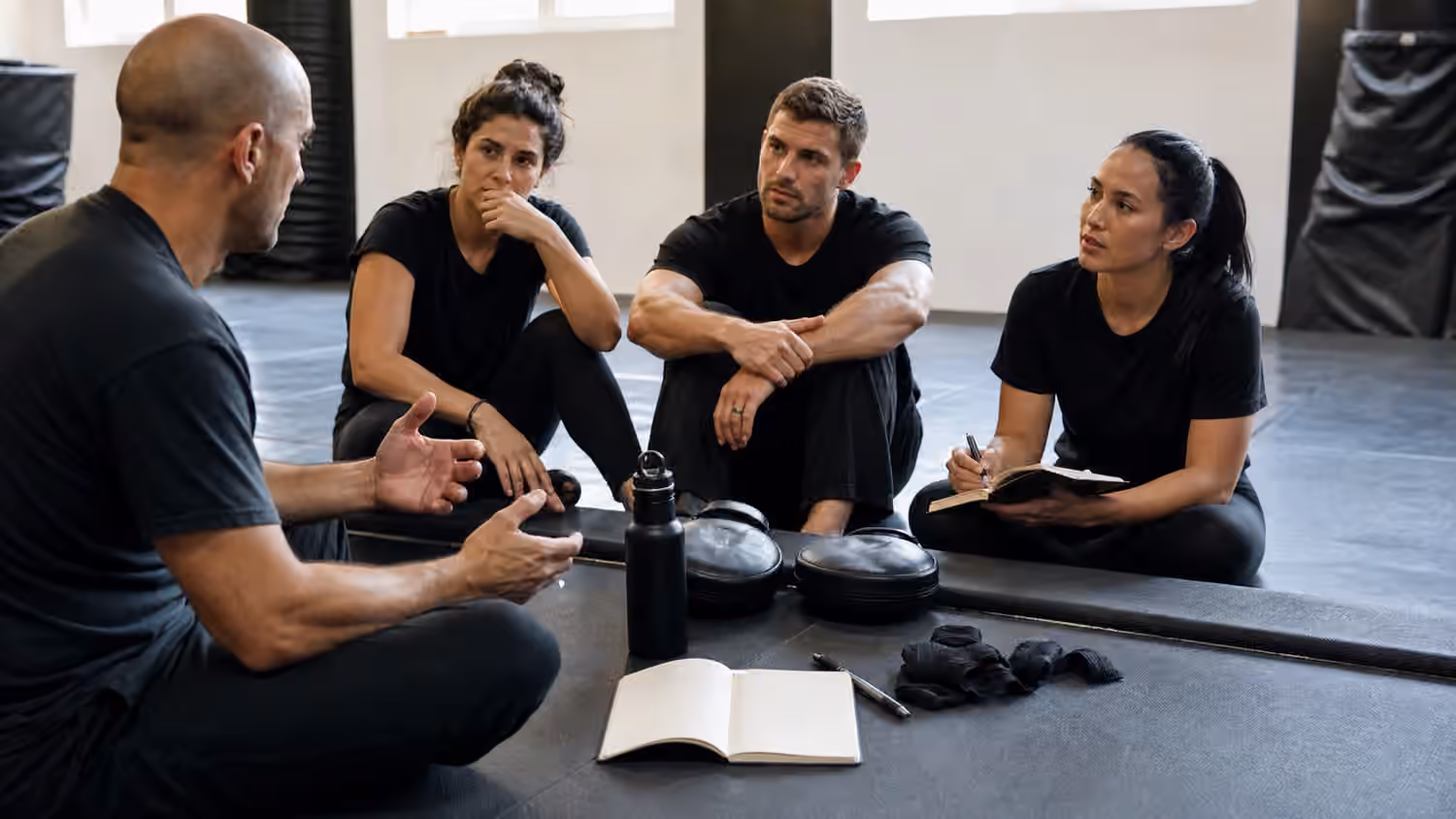 Krav Maga students sit on a training mat after class with pads, wraps, water, and a blank notebook.