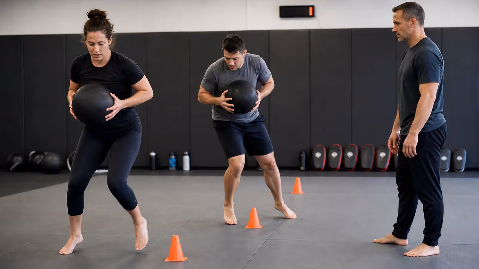 Adult Krav Maga students doing controlled conditioning drills with medicine balls and cones while a coach watches posture.