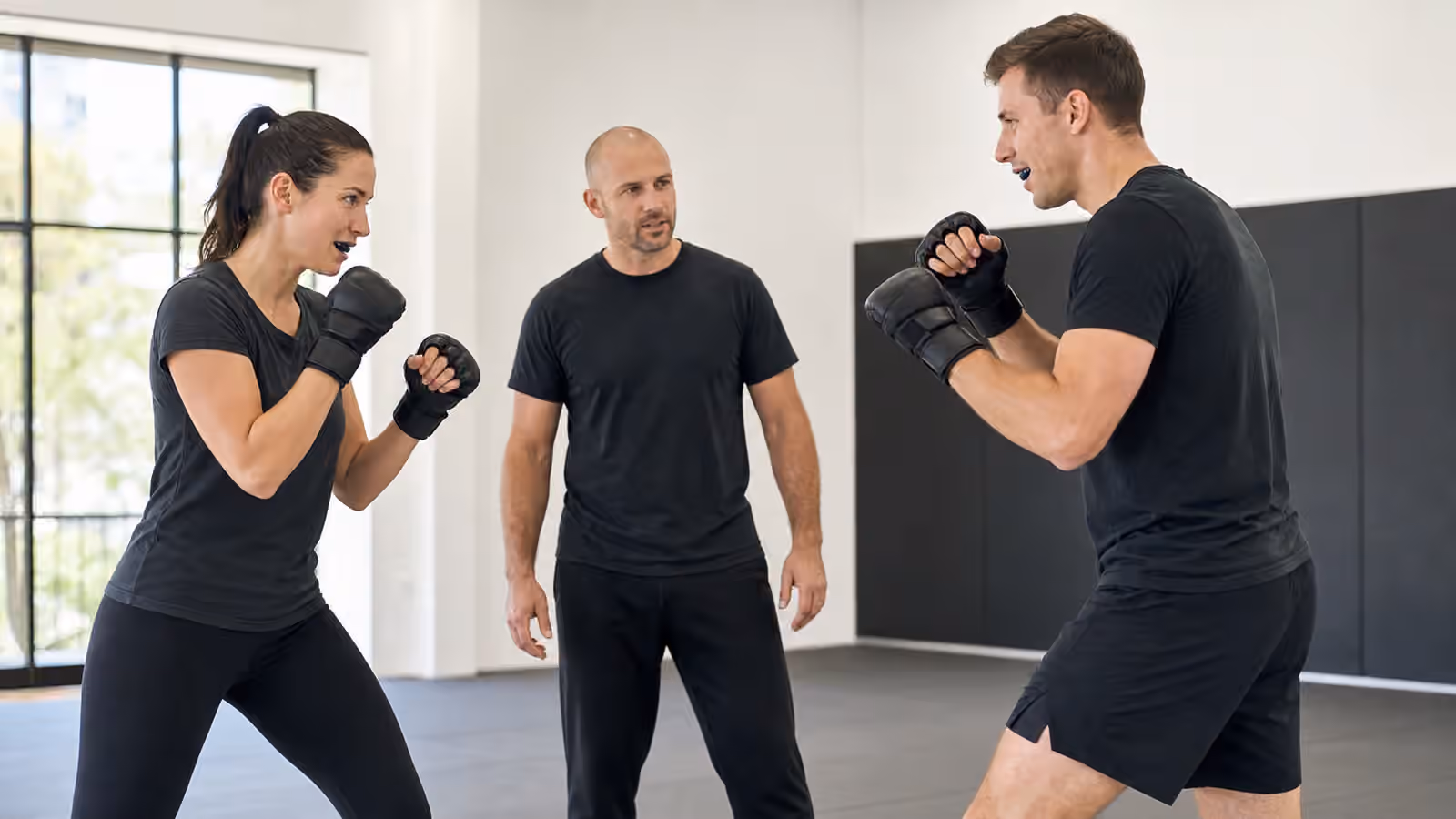 Two adult Krav Maga students practice controlled light sparring while an instructor observes spacing.