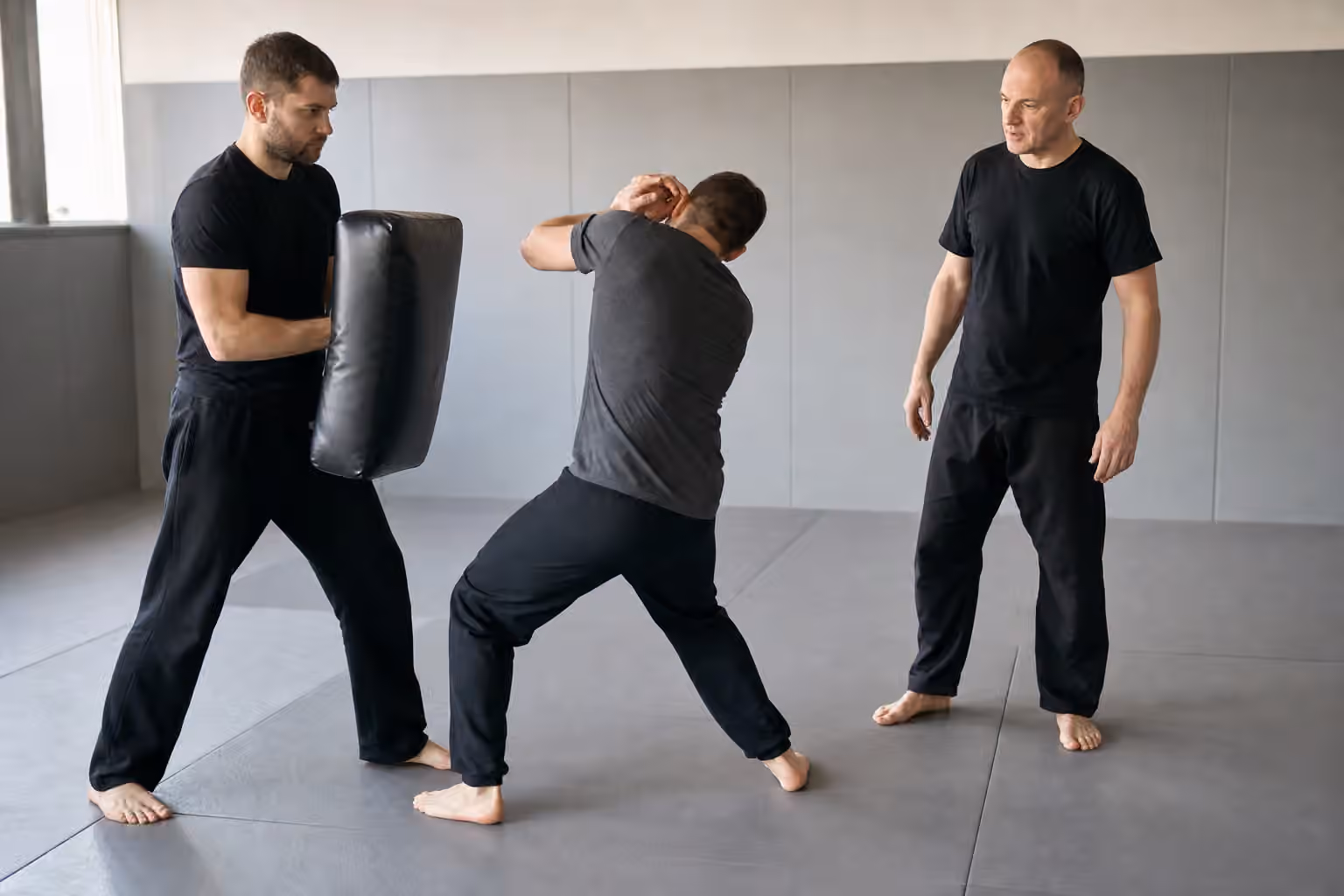 Adult Krav Maga students practicing a controlled head-protection cover and exit drill with a padded shield.