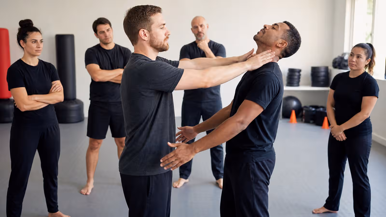 Adult Krav Maga students practice a slow neck-pressure safety drill while classmates and an instructor observe.
