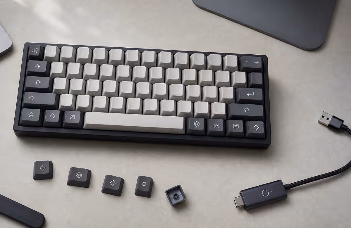 Compact mechanical keyboard with blank modifier keycaps, loose caps, USB adapter, and monitor base on a clean desk.