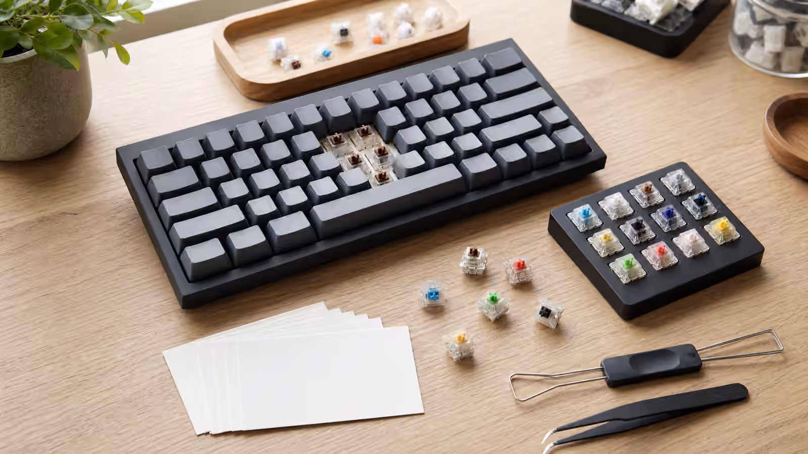 Mechanical keyboard with several switches removed, a switch tester, loose sample switches, blank note cards, and tools on a desk.