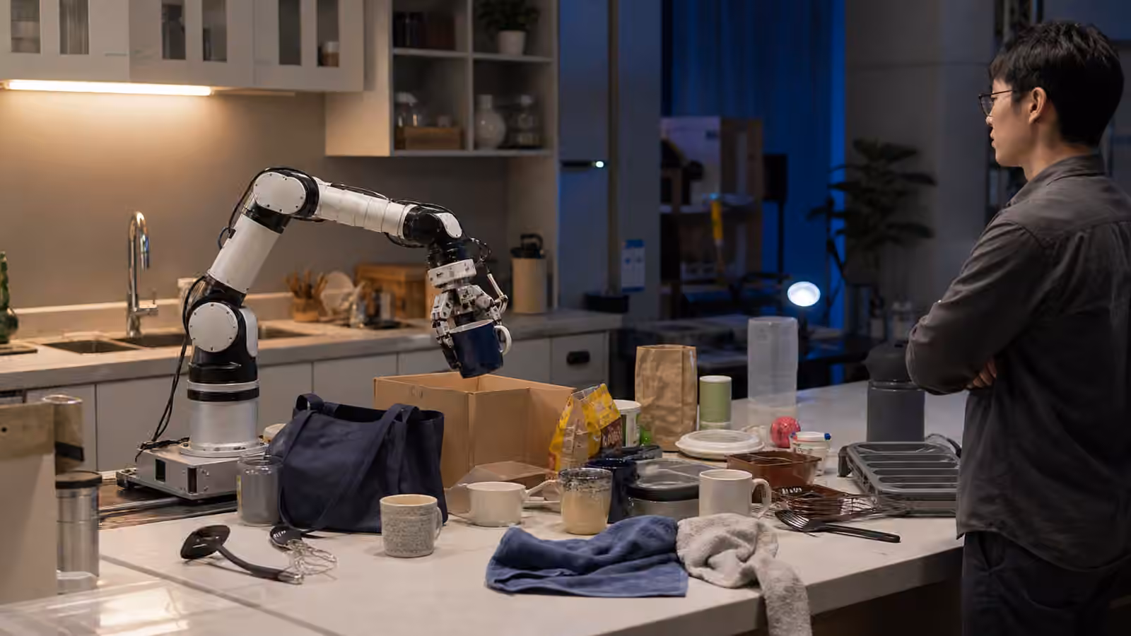 A robot arm practices ordinary kitchen object handling with mugs, towels, utensils, boxes, and clutter under varied lighting