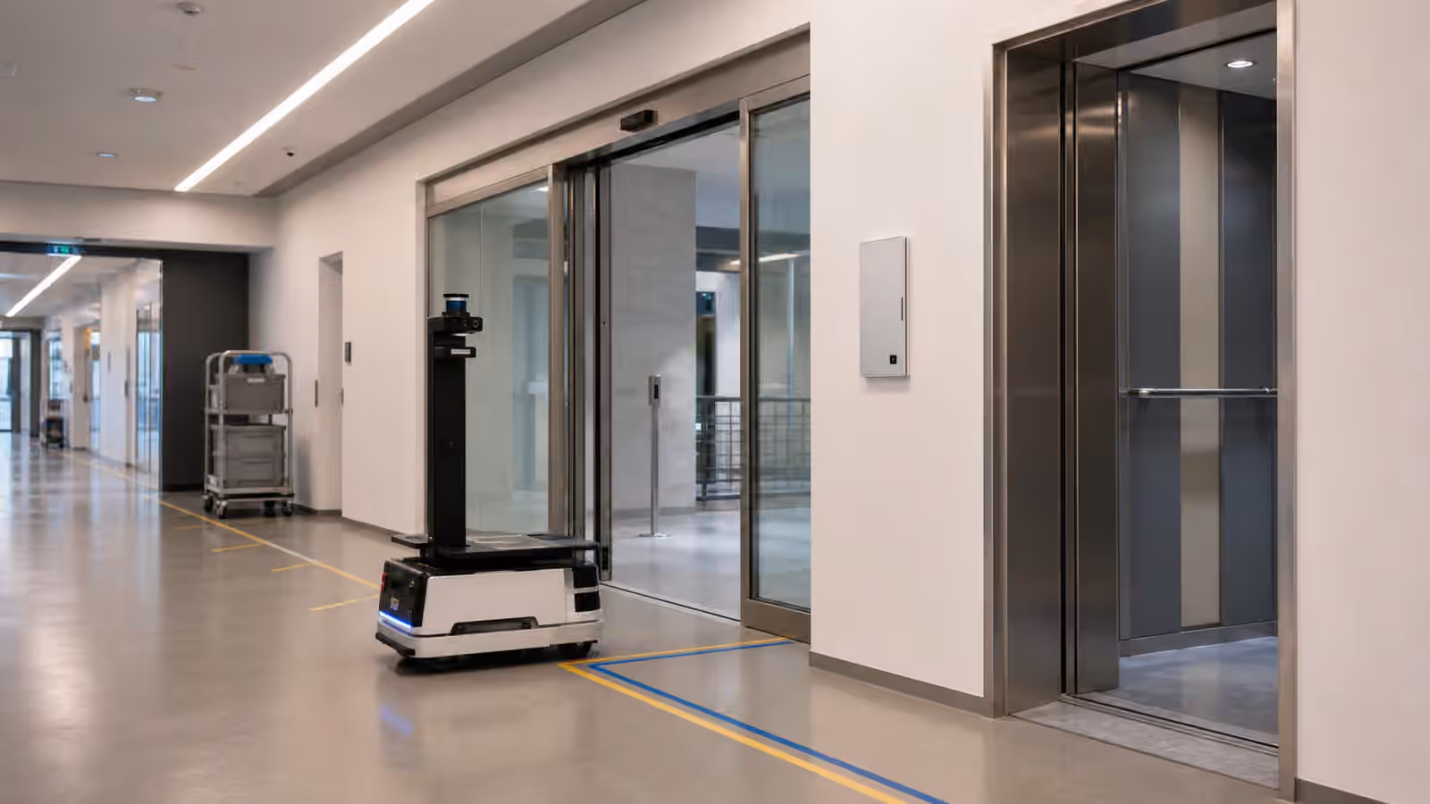 A mobile robot paused near an automatic door and elevator lobby in a modern facility corridor.