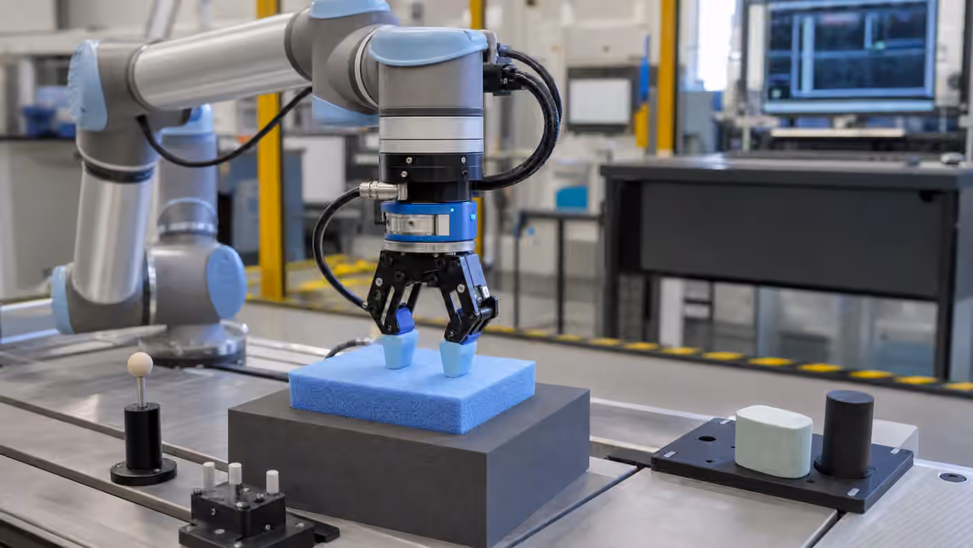 A robot arm on a lab bench gently pressing a compliant test block with force sensors, tactile pads, fixtures, safety tape, and an unreadable workstation nearby.