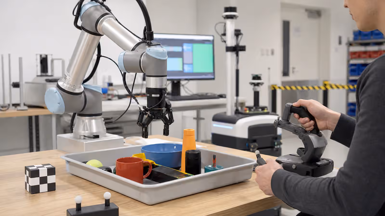 A collaborative robot arm in a lab learning from a human operator guiding a compact controller beside a tray of household objects.