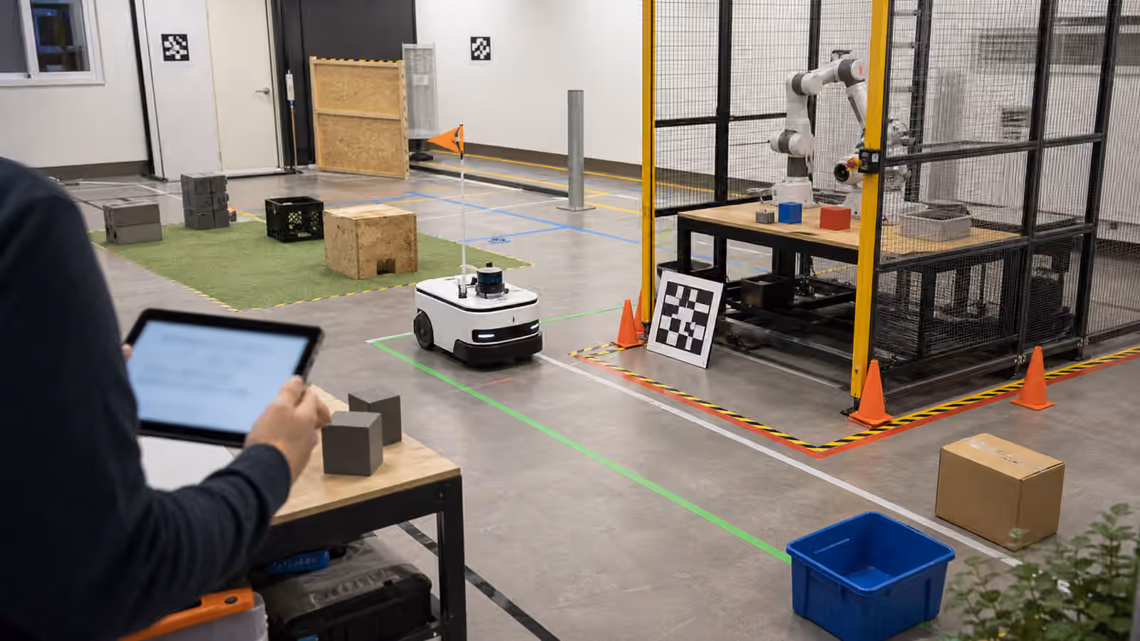 A mobile robot paused at the edge of a marked lab test lane beside a bounded robot arm workcell.