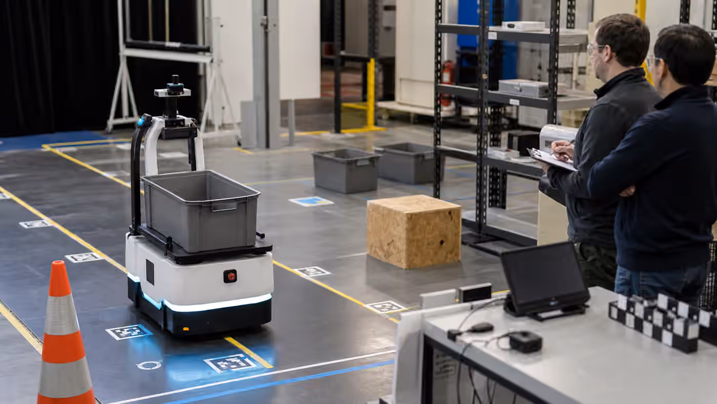 A mobile robot carrying a tote through a marked lab test lane while engineers observe with blank evaluation materials.