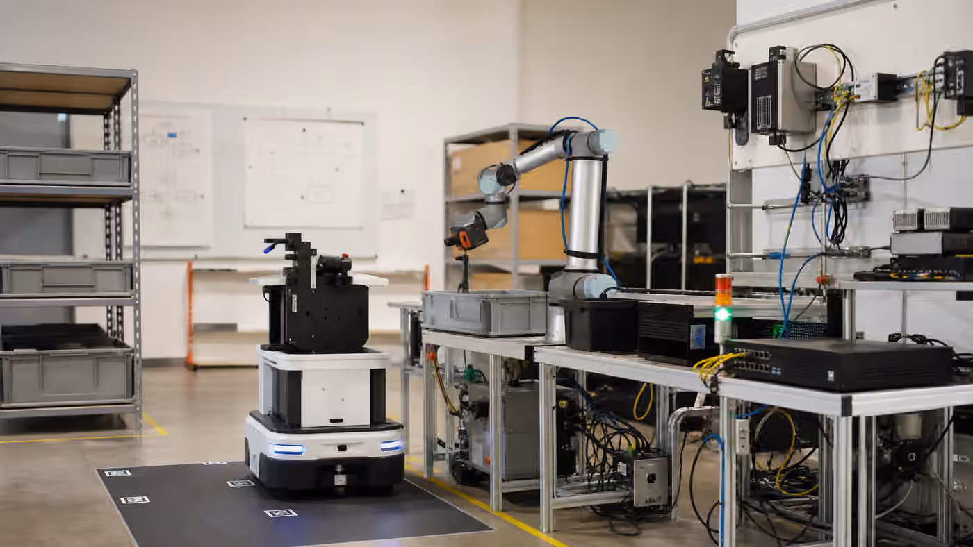 A mobile robot and robot arm in a lab surrounded by bins, network hardware, cables, and integration test equipment.