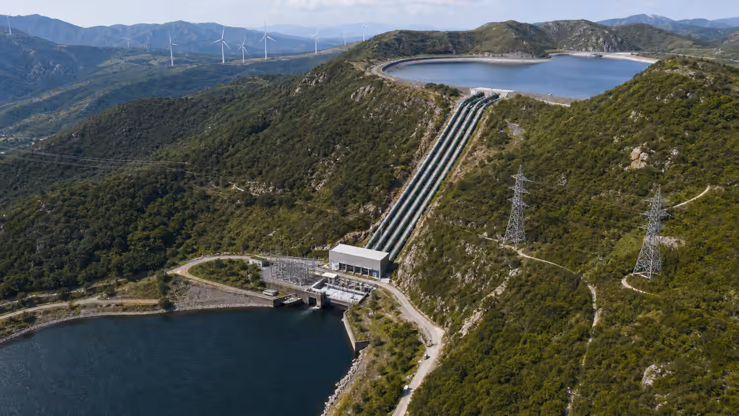 A pumped storage hydropower facility with an upper reservoir, lower reservoir, penstock pipes, a powerhouse, and transmission towers in hilly terrain.