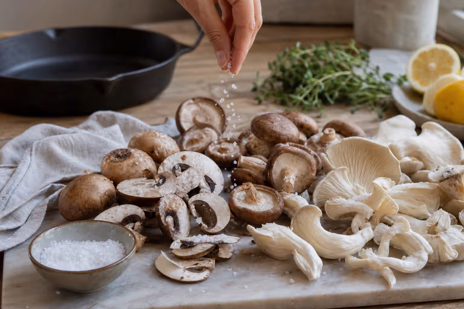 Mixed mushrooms being salted beside a cast iron skillet, coarse salt bowl, herbs, lemon, and a kitchen towel.