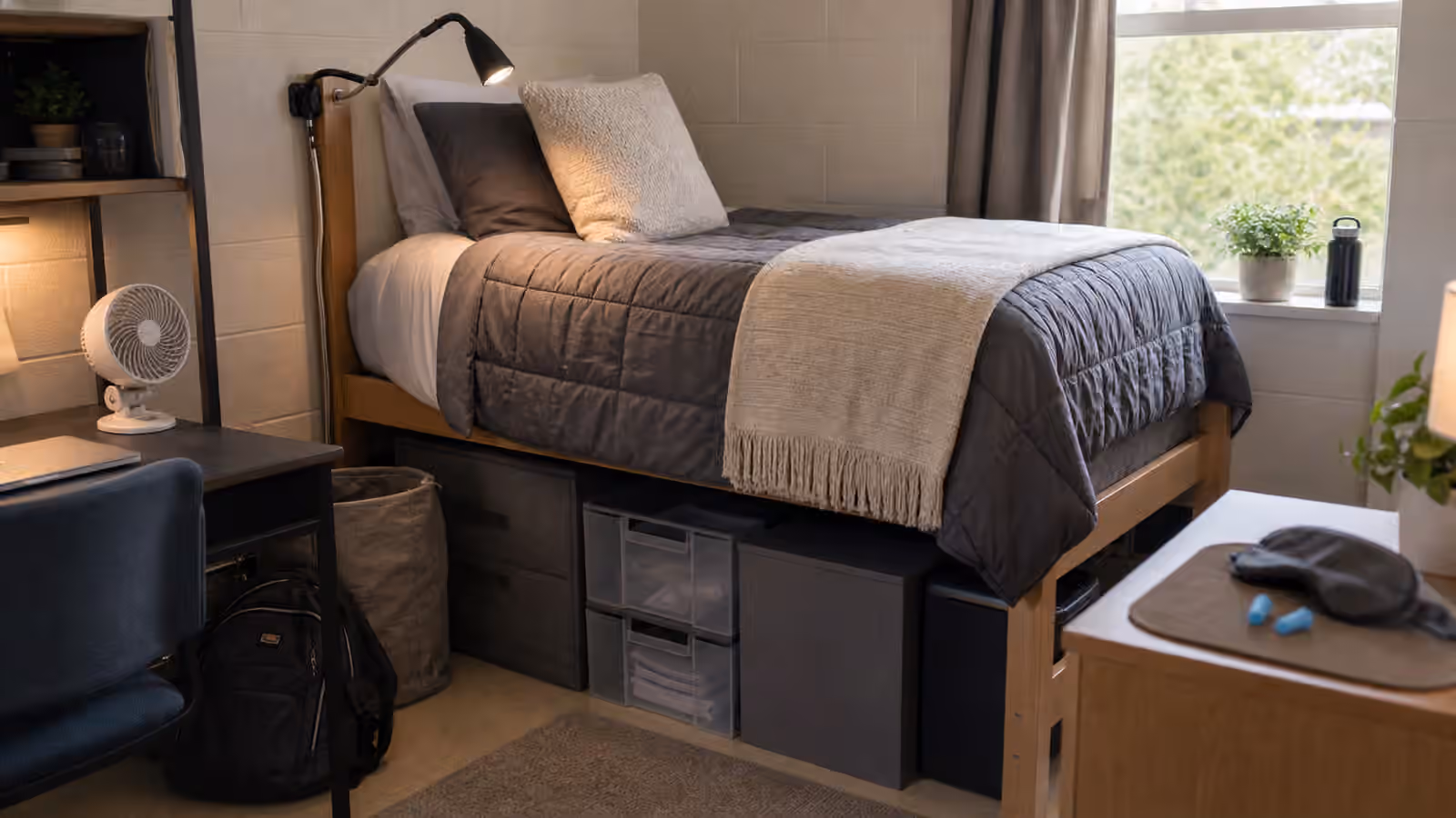 A compact dorm-style sleep setup with twin bed, under-bed storage, fan, lamp, and sleep mask tray.