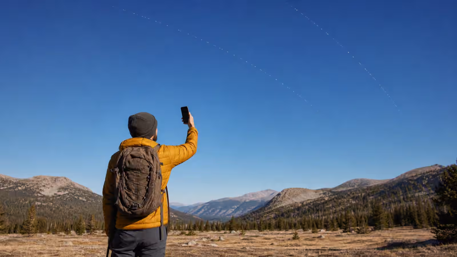 A hiker holds a phone toward open sky in a remote clearing while faint satellite tracks suggest a basic direct-to-phone link