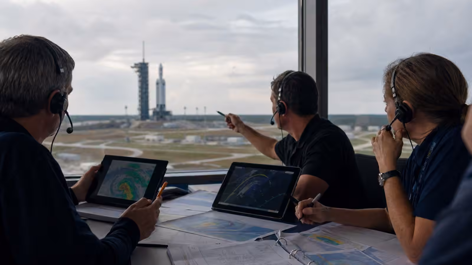 Launch planners compare weather, range, and orbital timing information while a rocket waits on a distant pad