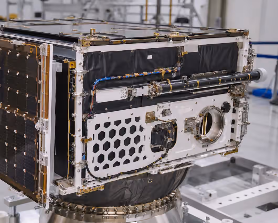 An unbranded satellite bus in a clean integration bay with structural panels, cable harnesses, folded deployable hardware, and a separation ring.