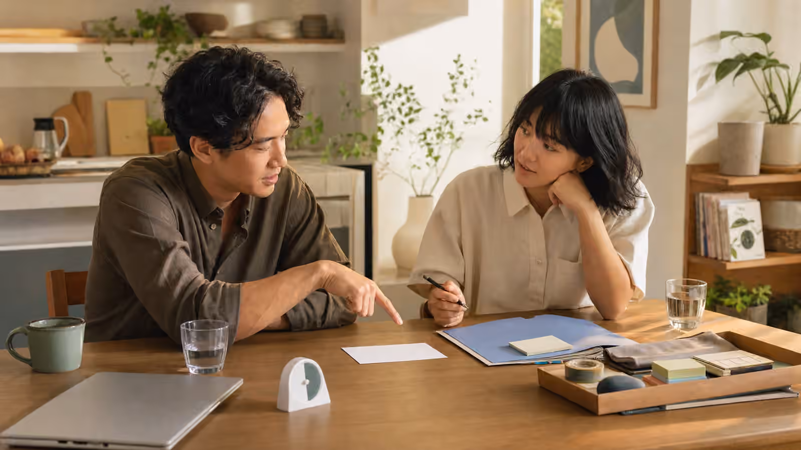 Two people at a shared table with a blank note card, folder, closed laptop, water glasses, timer, and task materials.