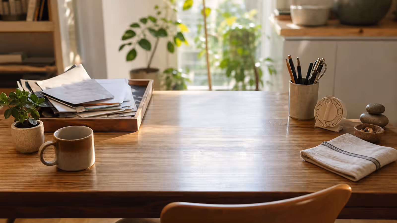 A warm table with one clear work area, a tray of papers, a timer, pens, cloth, mug, and grouped objects at the edge.