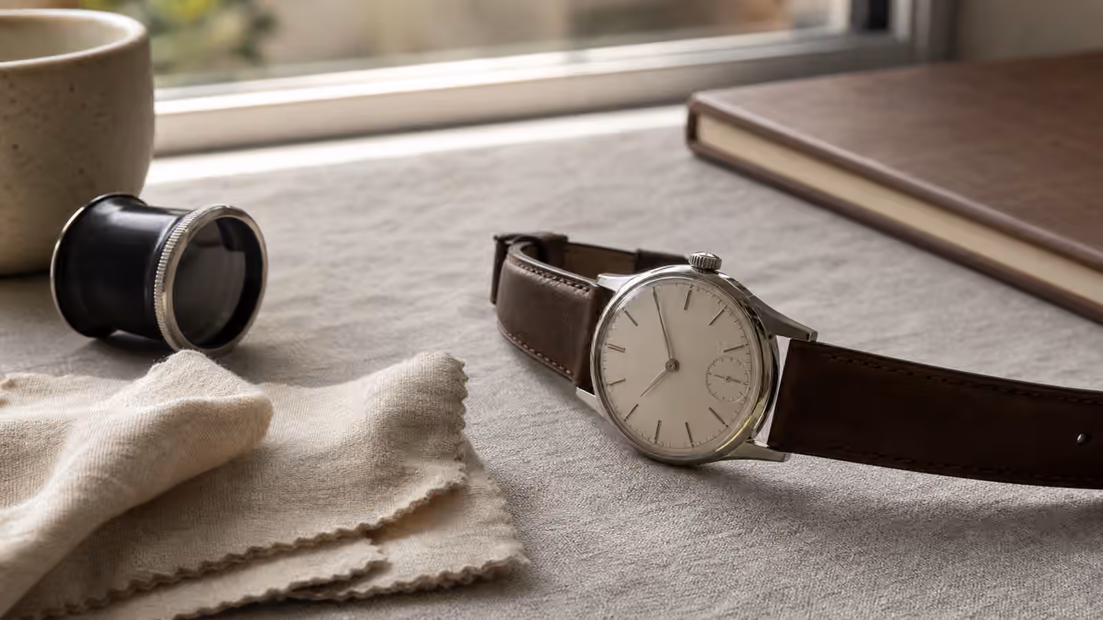 A slim unbranded hand-wound watch on a neutral cloth with a leather strap, loupe, and morning desk objects nearby.