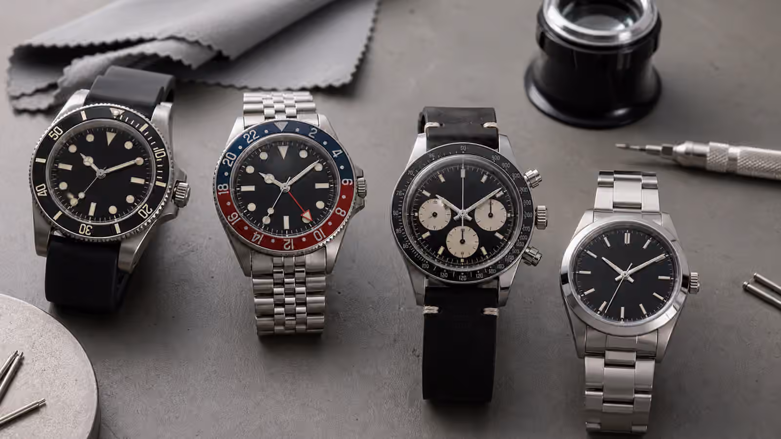 Unbranded watches with dive, GMT, chronograph, and fixed bezels rest on a gray watchmaker bench.