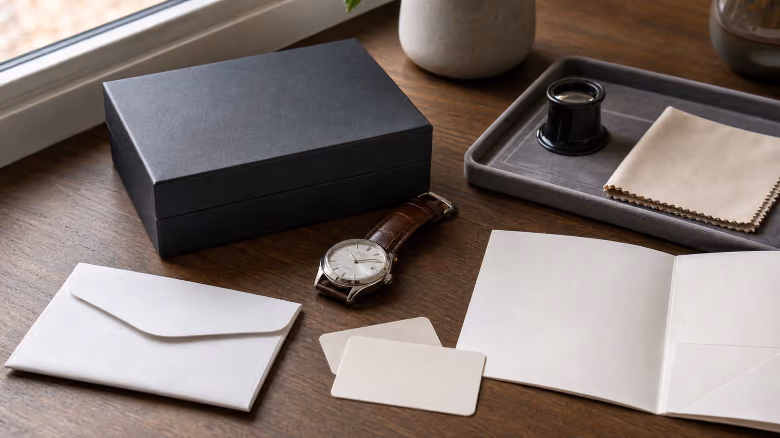 An unbranded watch rests on a wooden desk beside a plain box, blank cards, a service envelope, a loupe, and a cloth.