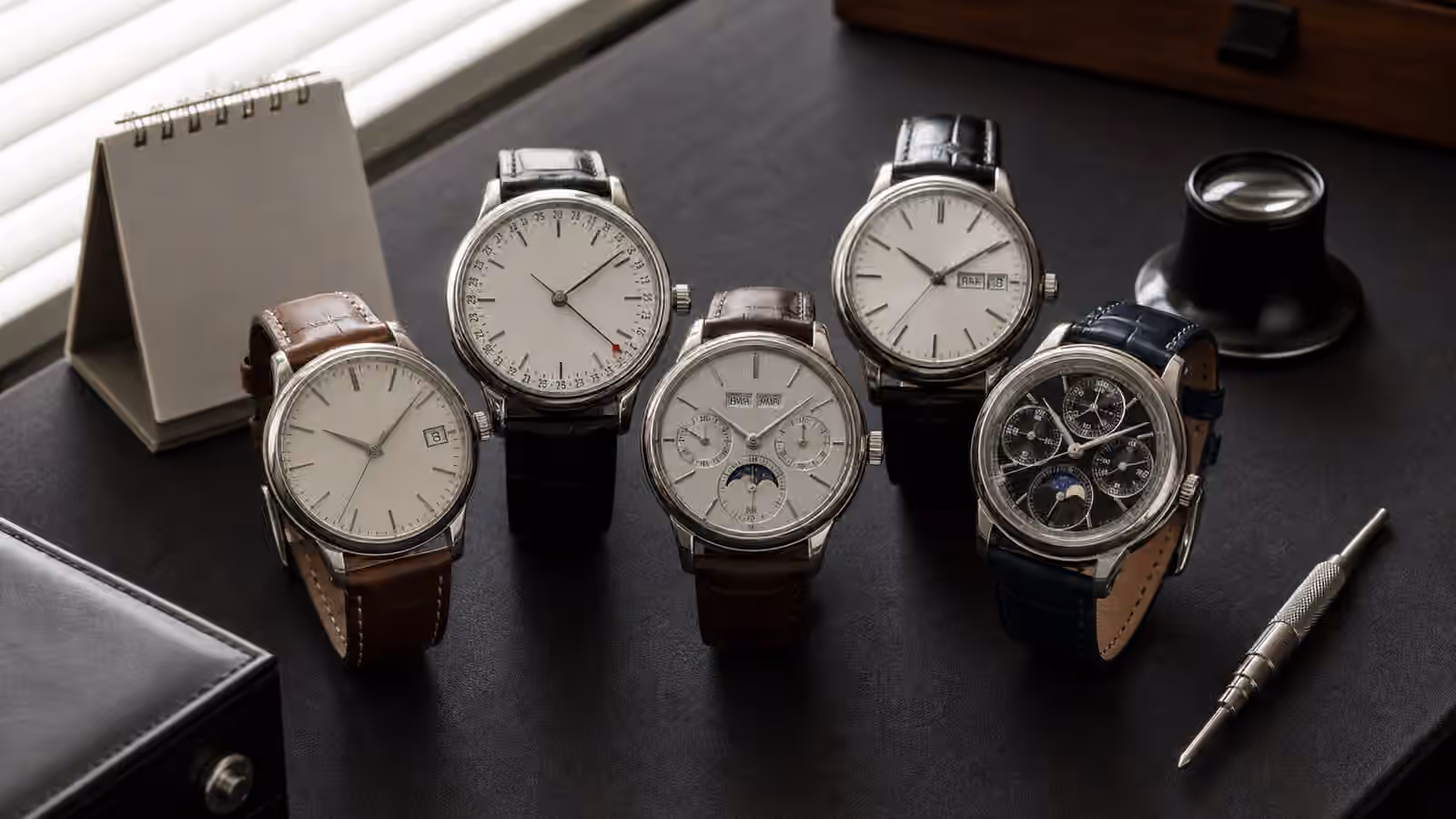 Several unbranded watches with calendar-style displays sit on a dark desk beside a loupe and blank calendar.