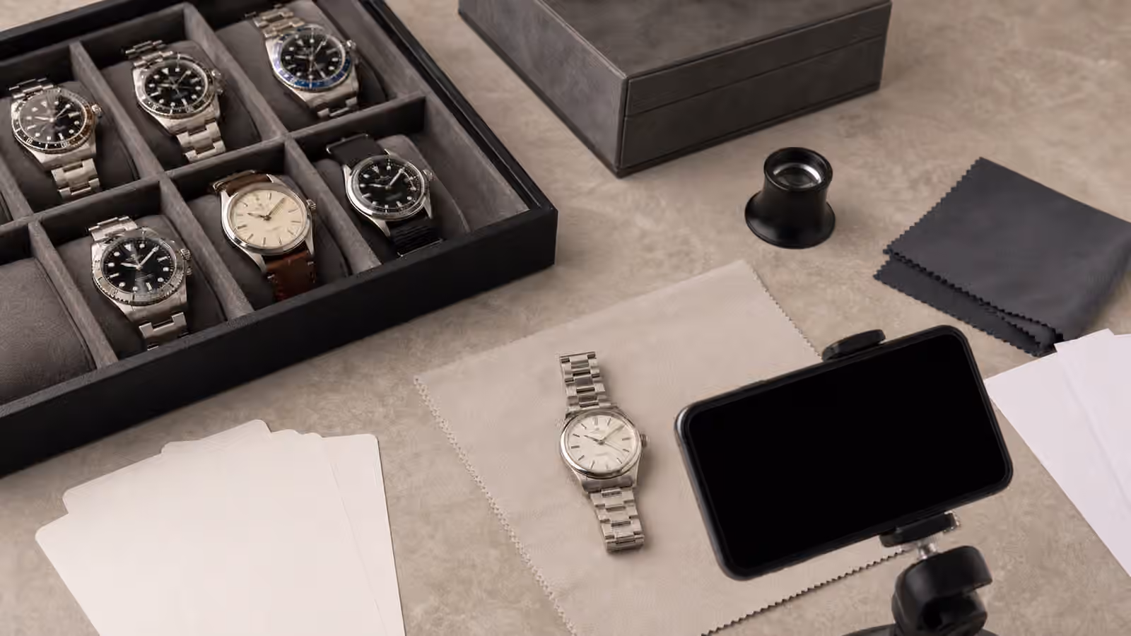 Several unbranded watches on a collector desk with a tray, blank cards, a loupe, a camera, a microfiber cloth, and storage papers turned face down.
