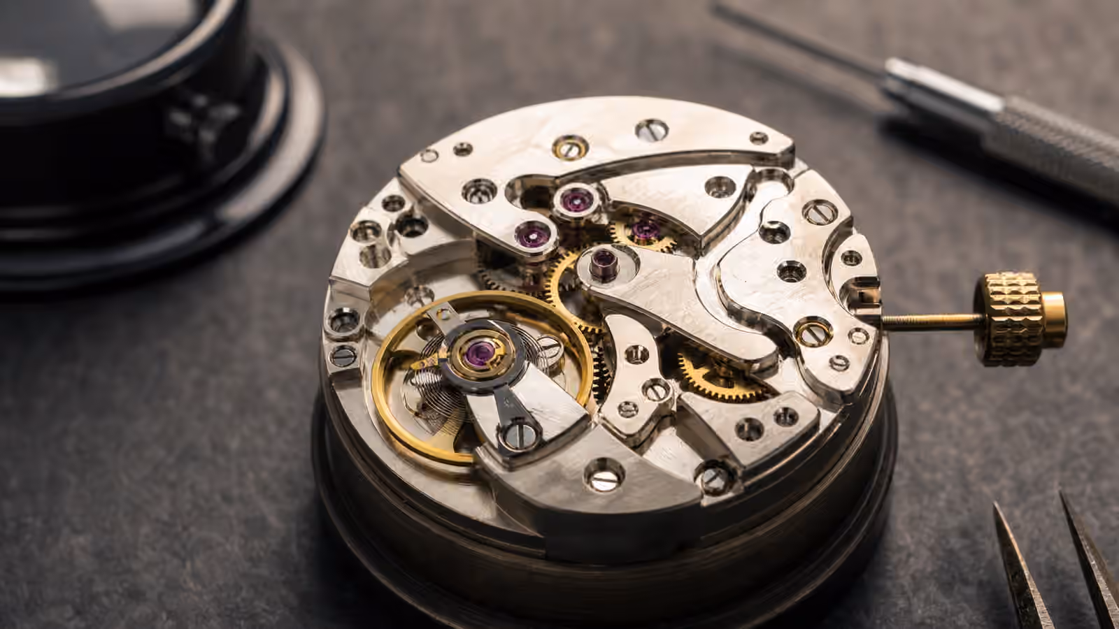A close view of an unbranded mechanical watch movement shows the balance wheel, escapement, bridges, screws, and jewel bearings on a watchmaker bench.