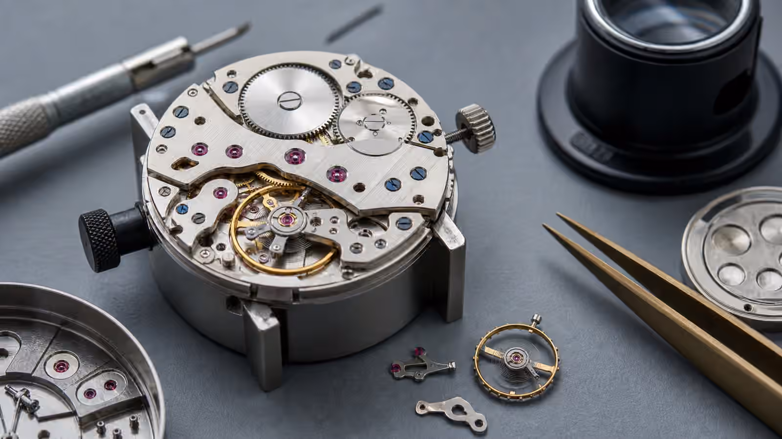 An unbranded mechanical watch movement on a gray work mat with ruby-colored jewels, gears, a balance wheel, tweezers, and a loupe.