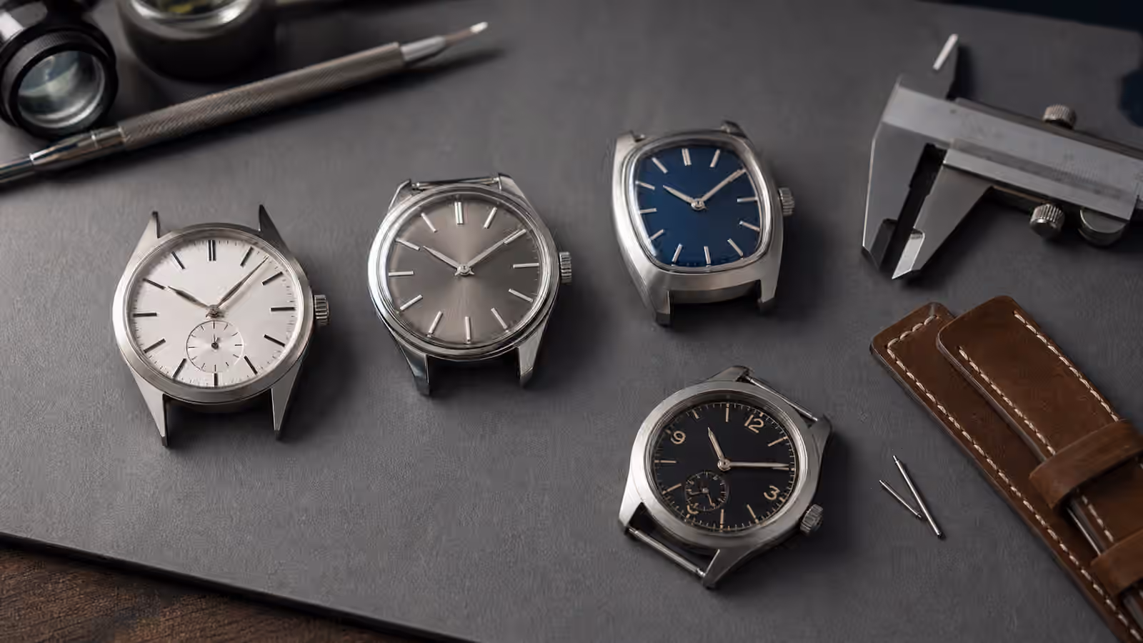 Unbranded watches with different lug shapes and case silhouettes rest on a gray watchmaker mat with calipers and straps nearby.