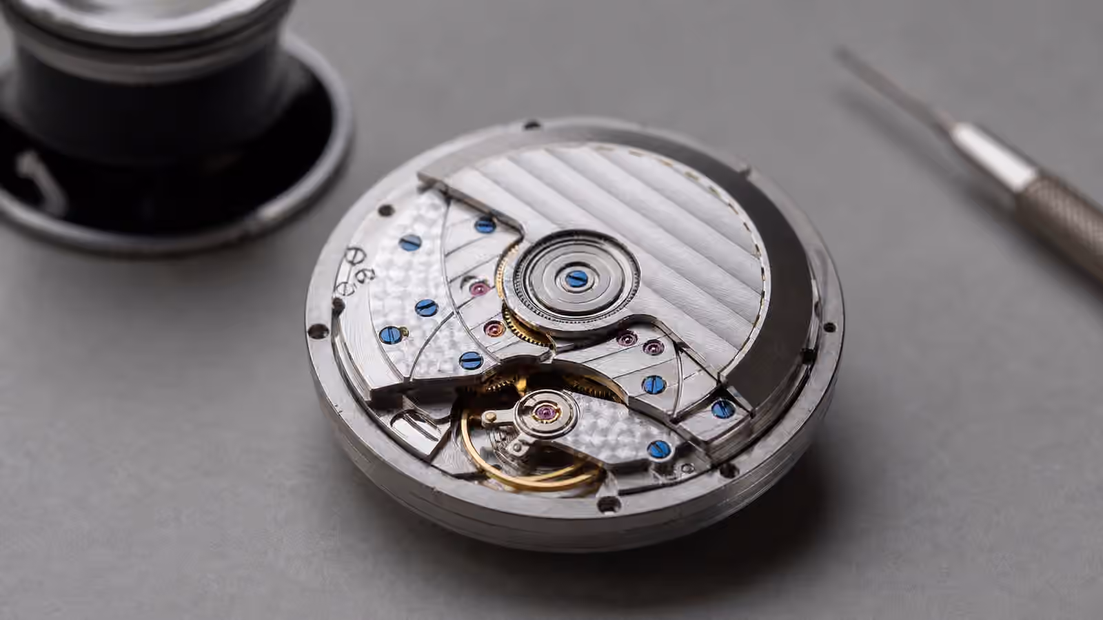 An unbranded mechanical watch movement on a gray watchmaker bench with visible striping, perlage, polished bevels, jewels, and blued screws.