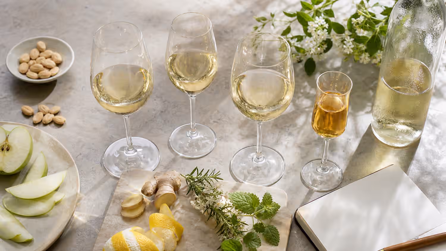 Aromatic white wine tasting flight with pale white wines, one amber-gold pour, citrus, apple, ginger, herbs, almonds, and a blank notebook.